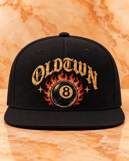 Black solid-back flat-bill OLDTWN snapback featuring gold OLDTWN lettering and a flaming eight-ball embroidered front design